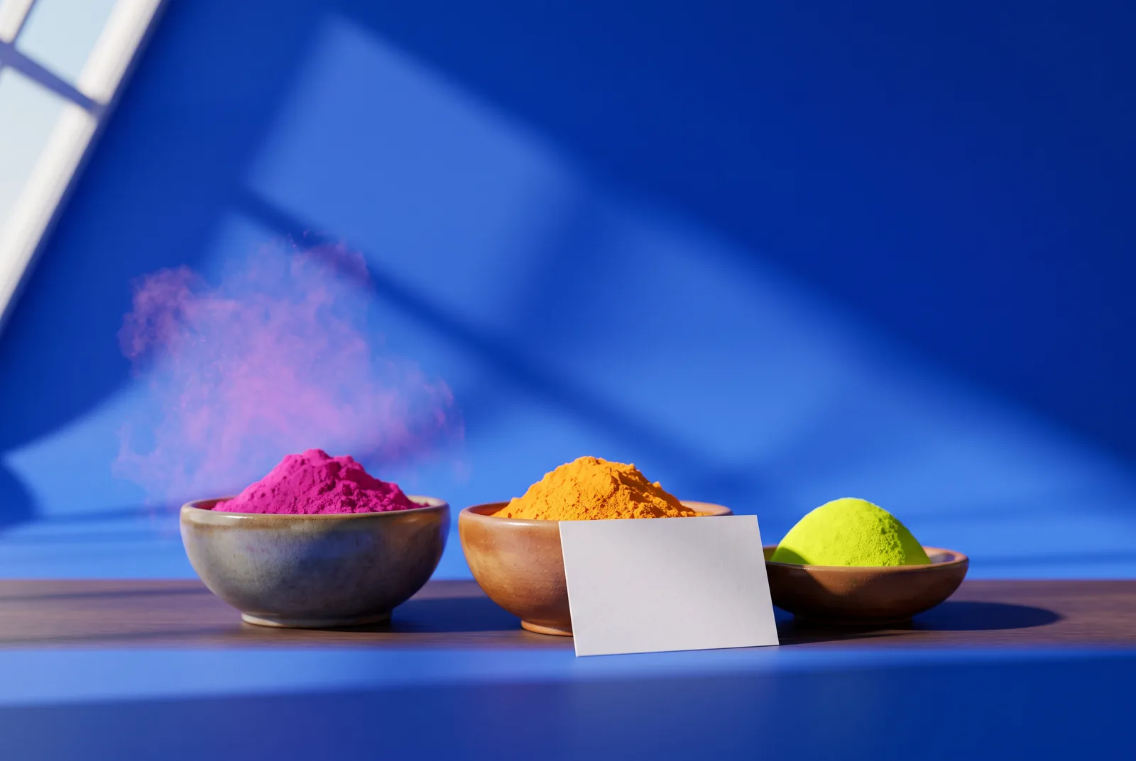 Holi campaign ideas: three bowls of bright color powder, magenta, saffron and lime green, with a puff of pink powder on a cobalt blue backdrop.