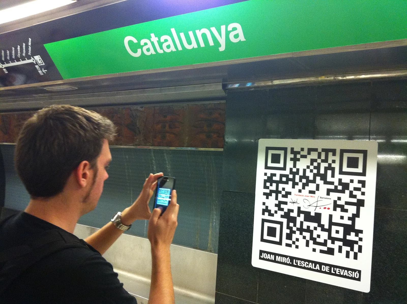 A man holding up a smartphone to scan a large QR code poster on a station wall in Barcelona.