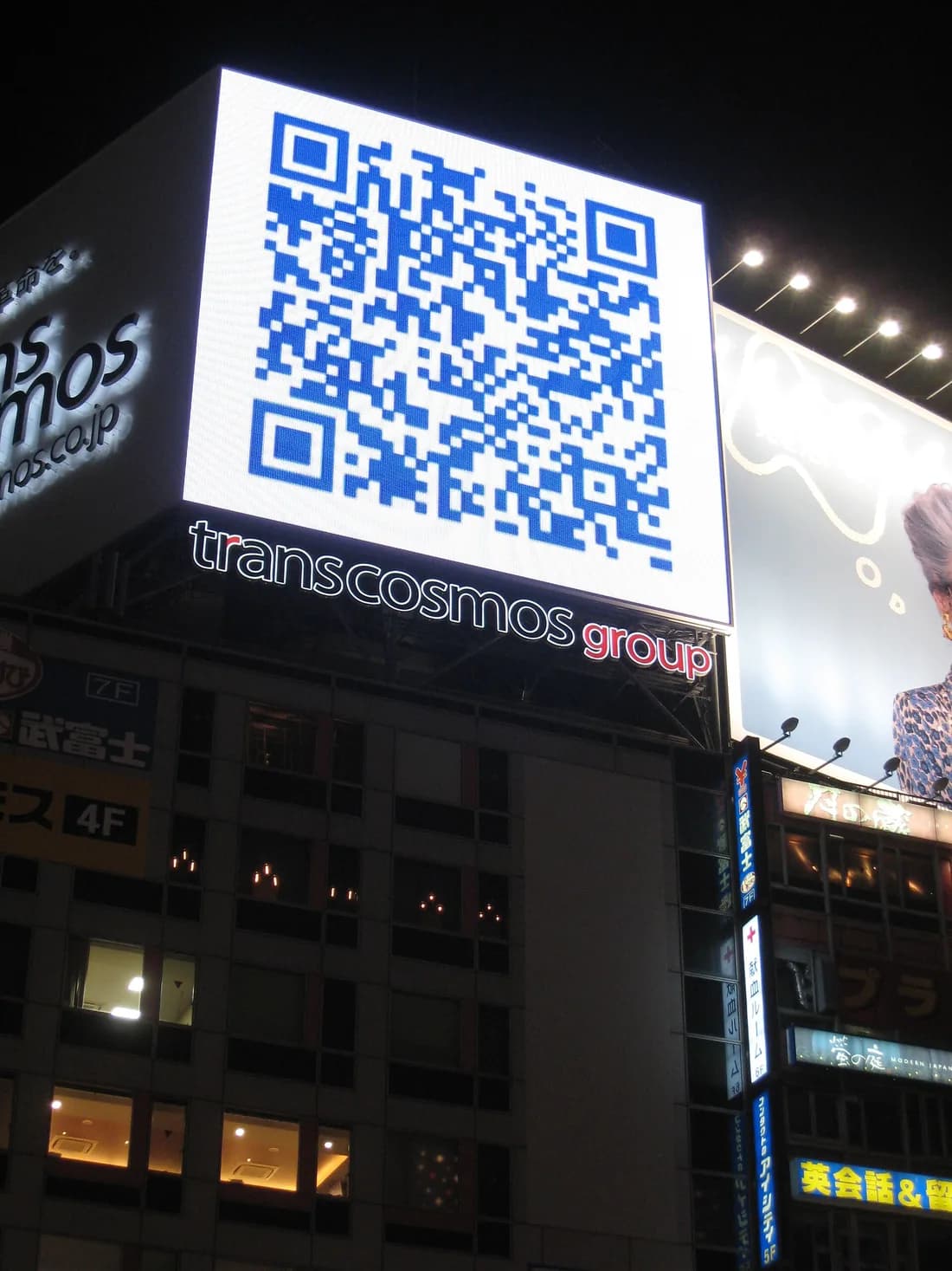 A giant blue QR code lit up on a billboard above a street in Shibuya, Tokyo, at night.