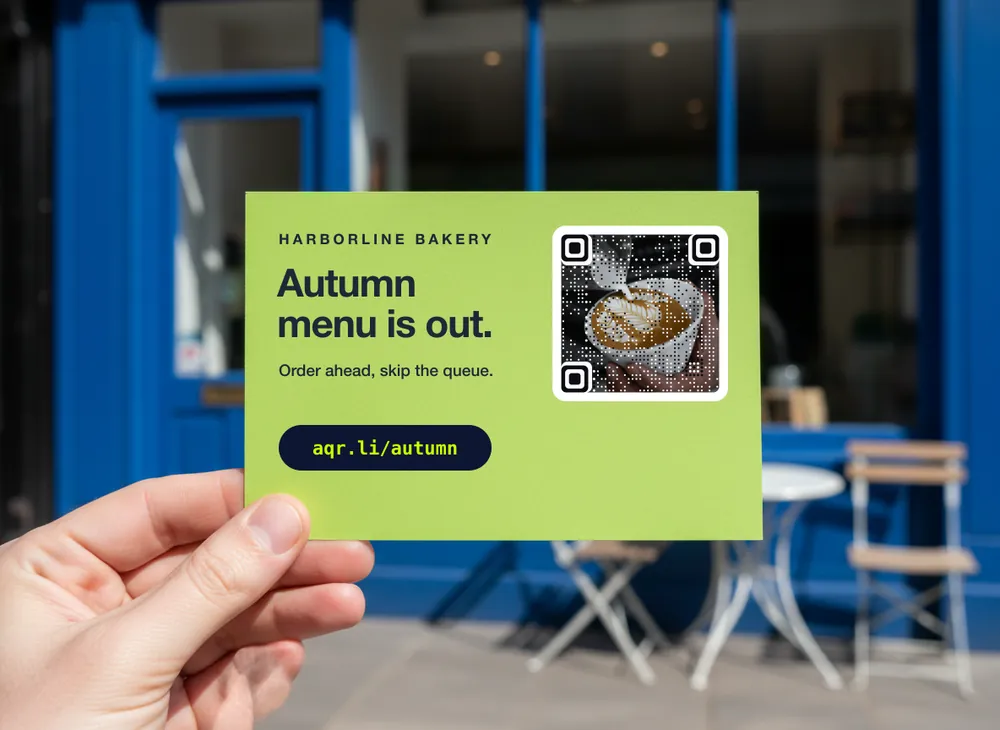 O mână ține un card lime tipărit, cu meniul de toamnă al unei brutării, în fața unei vitrine albastru cobalt; arată linkul scurt aqr.li/autumn lângă un cod artistic făcut dintr-o fotografie cu latte