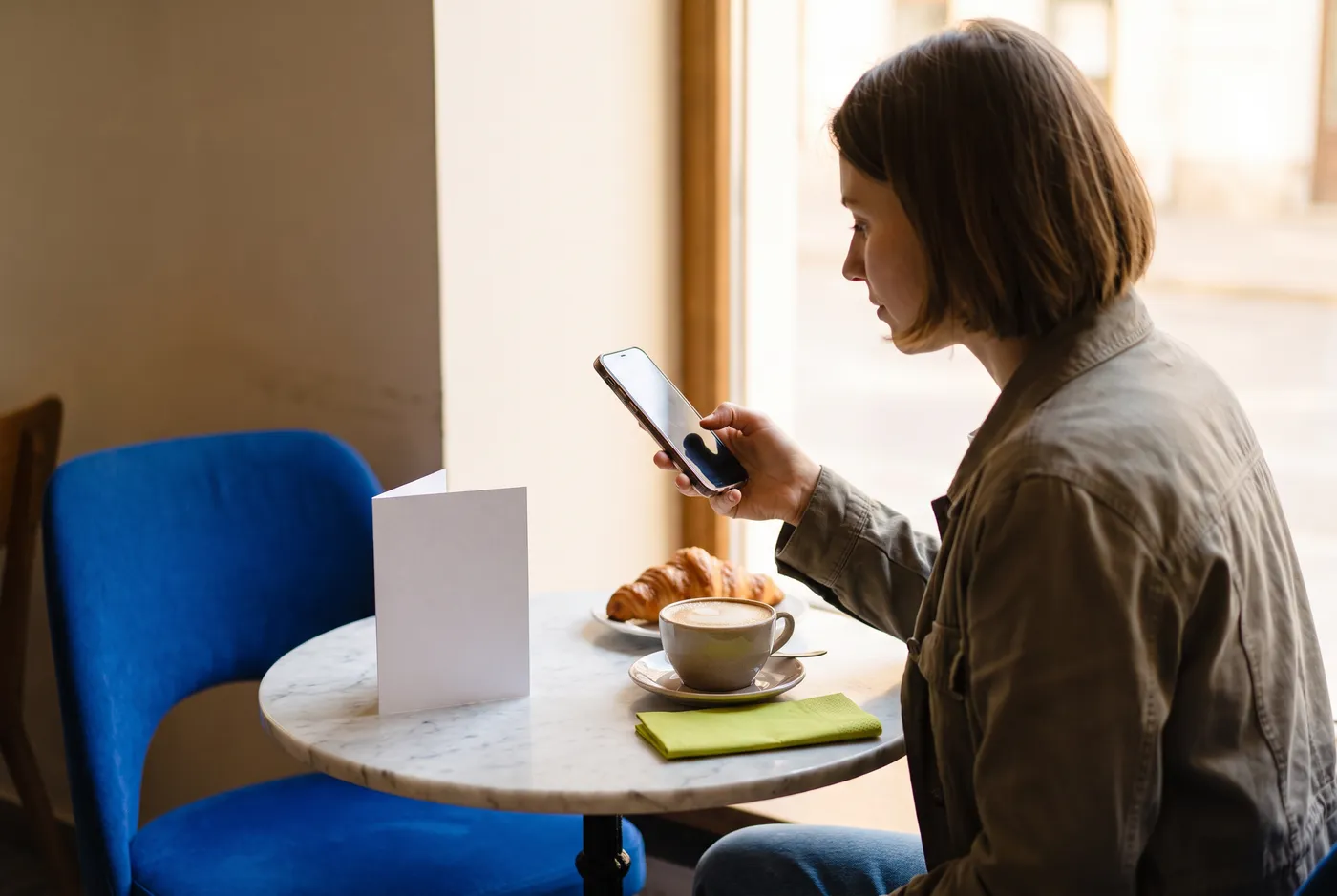 A customer at a café table holding her phone next to a small table card, with a cappuccino and a croissant, in warm afternoon light